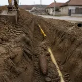 Un șanț lung săpat în pământ de-a lungul unei străzi rurale, cu o conductă galbenă de gaz vizibilă pe fundul acestuia
